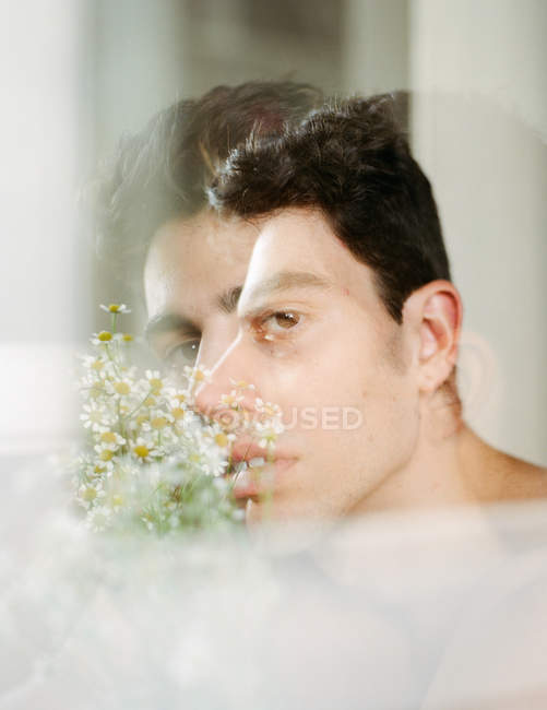 Vista laterale del giovane ragazzo senza maglietta con fiori bianchi freschi in bocca guardando la fotocamera su sfondo sfocato — Foto stock