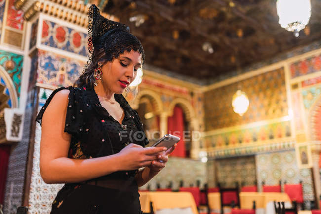 Vista lateral da jovem mulher no vestido usando telefone celular em pé perto da mesa com cadeiras no quarto de luxo do café decorado por mosaico — Fotografia de Stock