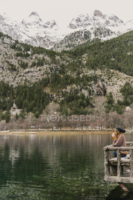 Seitenansicht einer Dame, die auf einer Bank sitzt und einen atemberaubenden Blick auf die Wasseroberfläche zwischen hohen Bergen mit Bäumen im Schnee und bewölkten Himmel in den Pyrenäen genießt — Stockfoto