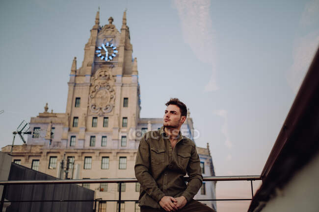 Giovane ragazzo elegante in abbigliamento casual vicino rotaie sul tetto guardando lontano vicino alla vecchia torre con orologi e cielo blu a Madrid, Spagna — Foto stock