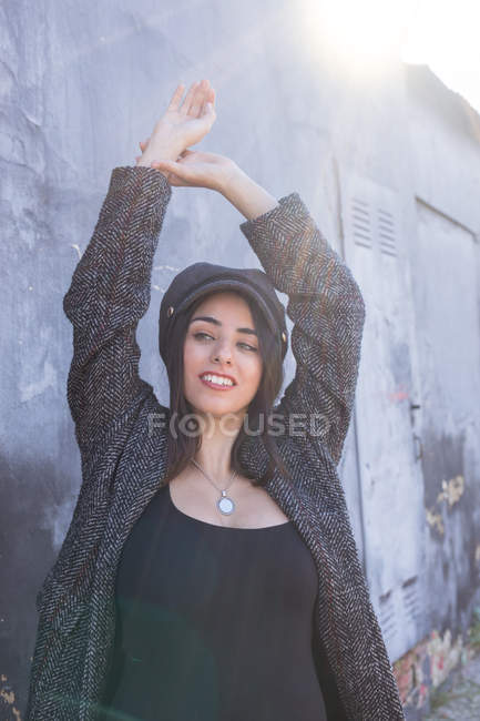 Mujer joven hispana sonriente con abrigo y gorra de pie cerca de la pared de mala calidad en retroiluminación - foto de stock