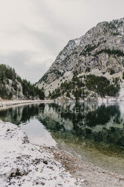 Increíble vista de la superficie del agua entre altas montañas con árboles en la nieve y el cielo nublado en los Pirineos - foto de stock