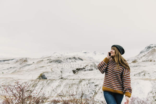 Jovencita sonriente en suéter y sombrero hablando por teléfono móvil y mirando lejos cerca de las colinas en la nieve en los Pirineos - foto de stock