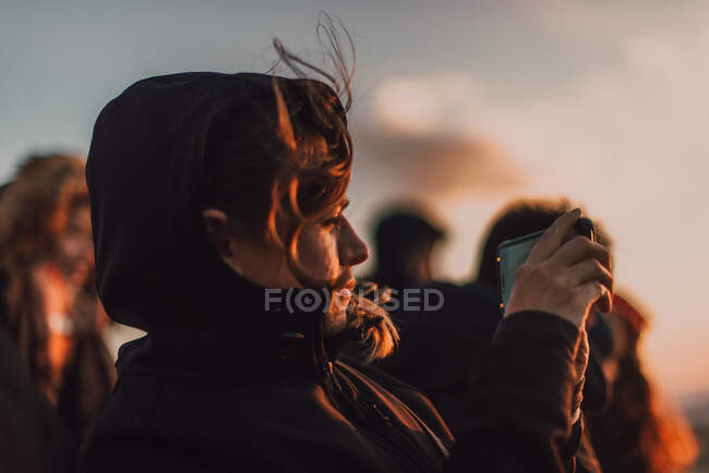 Vista lateral de la señora de abrigo tomando fotos en el teléfono móvil al atardecer en Chefchaouen, Marruecos - foto de stock