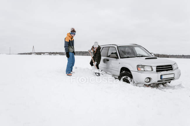 Женщина с лопатой рядом с мужчиной и машиной на снежном поле — стоковое фото