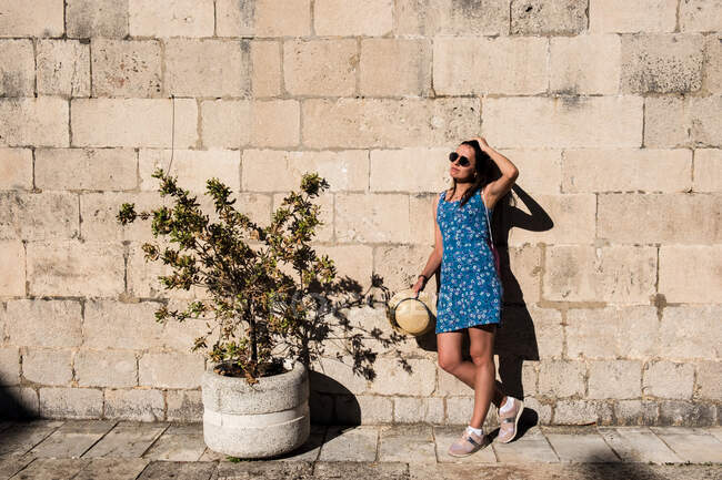 Attraktive junge Frau mit Sonnenbrille steht an einem sonnigen Tag in der Nähe einer alten Steinmauer und einer hübschen Topfpflanze — Stockfoto