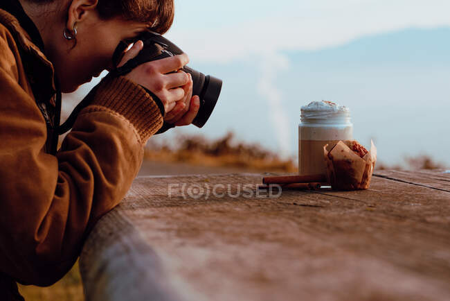Side view of young female in casual outfit using professional camera to take picture of aromatic coffee and delicious muffin while sitting at timber table in countryside — Stock Photo