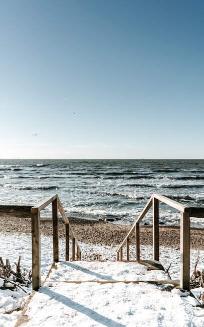 Escalones de madera en la nieve cerca de la costa del mar y el cielo azul en Klaipeda, Lituania - foto de stock