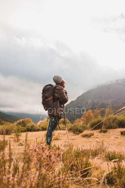 Seitenansicht einer Dame mit Hut und Skijacke mit Ranzen und Spazierstock, die auf einer Wiese in der Nähe eines Berges in Wolken in Isoba, Kastilien und León, Spanien, spaziert — Stockfoto