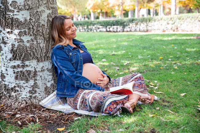 Улыбающаяся беременная привлекательная женщина сидит под деревом — стоковое фото