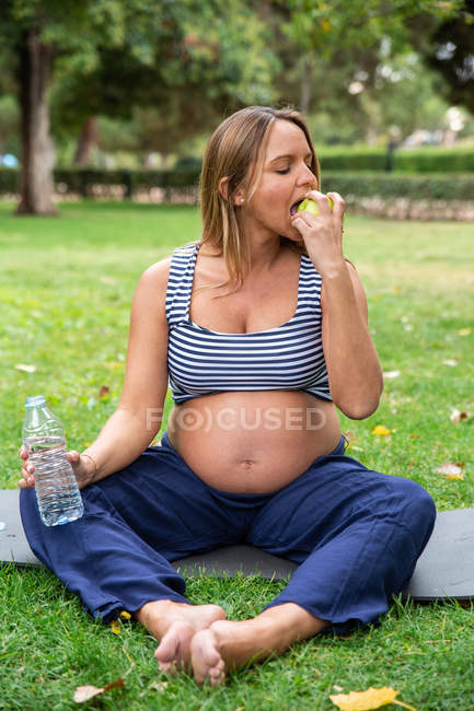 Young pregnant woman in sportswear holding bottle of water and eating fresh apple on yoga mat in green garden — Stock Photo