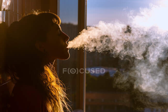Vista lateral da jovem com os olhos fechados fumando perto da janela no apartamento à noite — Fotografia de Stock
