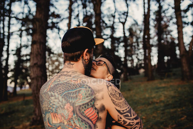 Вид сзади мужчины без рубашки с татуировками, обнимающего женщину в парке на размытом фоне — стоковое фото
