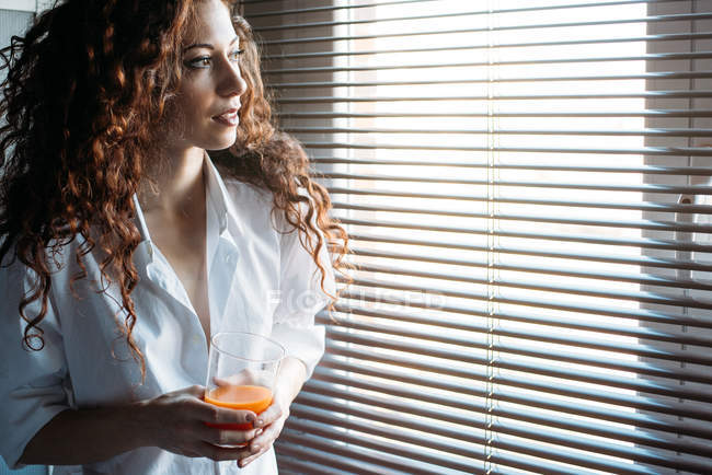 Hermosa mujer de pie junto a una ventana ciega bebiendo jugo de naranja - foto de stock