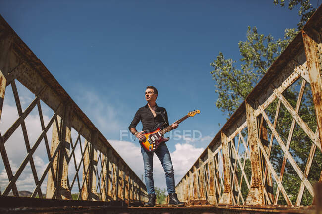 Ragazzo adulto con chitarra elettrica in piedi sul ponte intemperie e guardando altrove nella giornata di sole in campagna — Foto stock