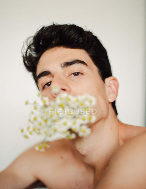 Боковой вид молодого парня без рубашки со свежими белыми цветами во рту, смотрящего в камеру на размытом фоне — стоковое фото