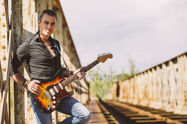 Adulte guy avec guitare électrique debout sur weathered pont et regarder caméra par jour ensoleillé dans la campagne — Photo de stock