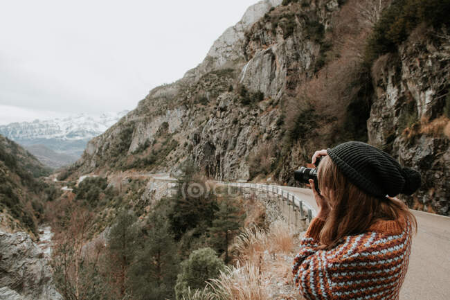 Vista lateral de la joven dama en suéter y sombrero tomando fotos en cámara entre colinas en los Pirineos - foto de stock