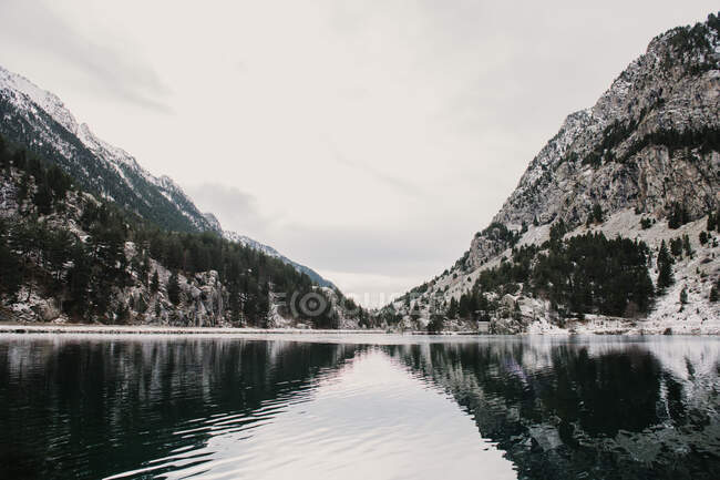 Increíble vista de la superficie del agua entre altas montañas con árboles en la nieve y el cielo nublado en los Pirineos - foto de stock