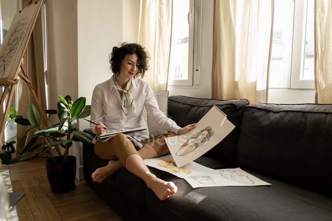 Jeune femme heureuse attrayante assise sur un canapé avec un ensemble de peintures sur des feuilles dans la pièce lumineuse — Photo de stock