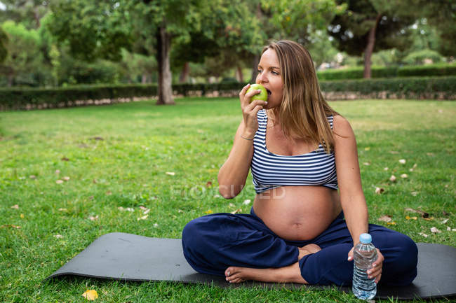 Беременная привлекательная женщина с водой и яблоком на коврике в парке — стоковое фото