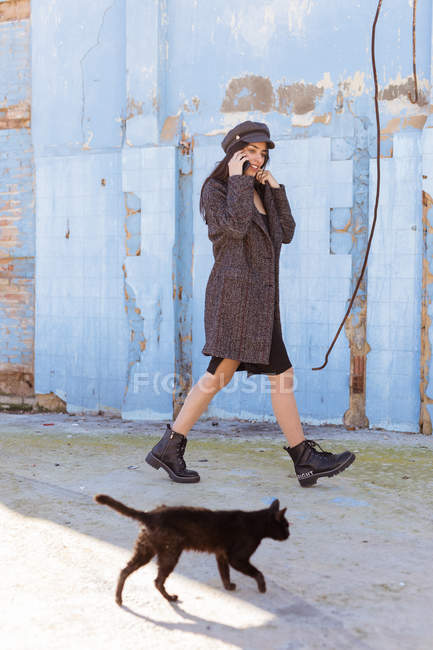 Elegante mujer joven con abrigo y gorra hablando en el teléfono móvil mientras camina cerca de la pared de ladrillo azul y gato negro - foto de stock