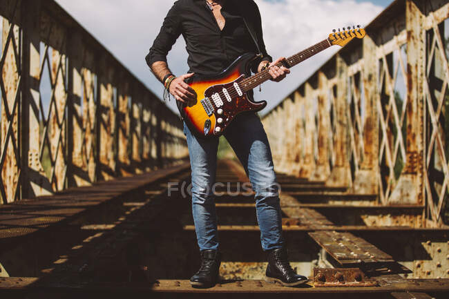Colheita Adulto cara com guitarra elétrica de pé na ponte weathered no dia ensolarado no campo — Fotografia de Stock