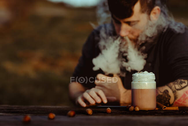 Mann dampft und benutzt Smartphone in der Nähe einer Tasse frischen Kaffees, während er am Holztisch im Grünen sitzt — Stockfoto