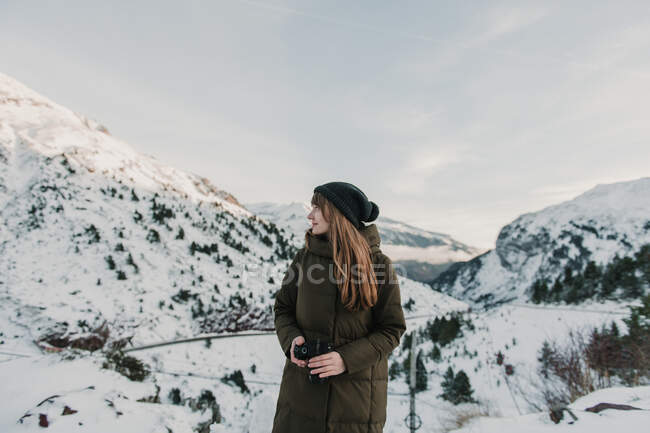 Jeune femme en veste de ski et chapeau tenant la caméra et regardant loin entre les collines dans la neige dans les Pyrénées — Photo de stock
