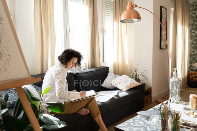 Seitenansicht einer jungen attraktiven Frau, die auf einem Sofa sitzt und auf einem Laken in der Nähe eines niedrigen Tisches mit Pinseln und Bleistiften in Dosen schreibt — Stockfoto