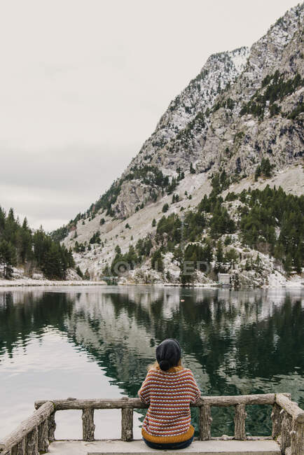 На задньому плані жінка сидить на лавці і дивиться на дивовижну поверхню води між високими горами з деревами в снігу і хмарному небі в Піренеях. — стокове фото