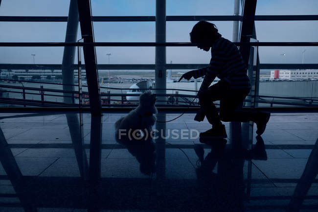 Silhueta de menino irreconhecível formação pequeno cão dentro escuro aeroporto edifício — Fotografia de Stock