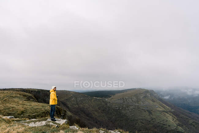 Side view of guy in yellow coat and hat standing on top of mountain and cloudy sky in Orduna, Spain — Foto stock