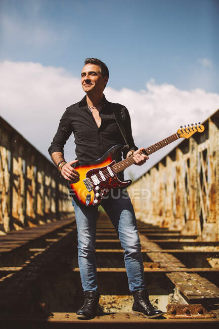 Adulte guy avec guitare électrique debout sur weathered pont et regarder loin par jour ensoleillé dans campagne — Photo de stock