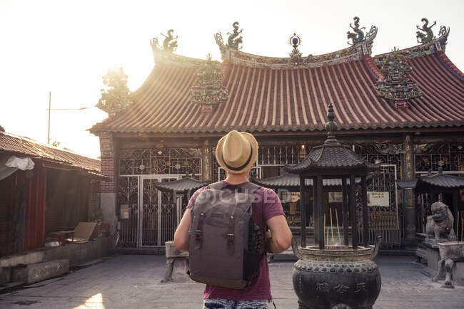 Вид сзади молодой парень в шляпе с рюкзаком, стоящим между азиатскими магазинчиками, старыми постройками и памятниками в Малайзии — стоковое фото