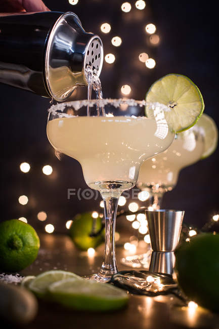 Pouring margarita cocktail in glasses from cocktail shaker on dark background with lights — Stock Photo