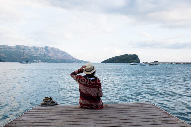 Back view of young female in stylish hat and patterned sweater sitting on pier and admiring view of beautiful sea - foto de stock