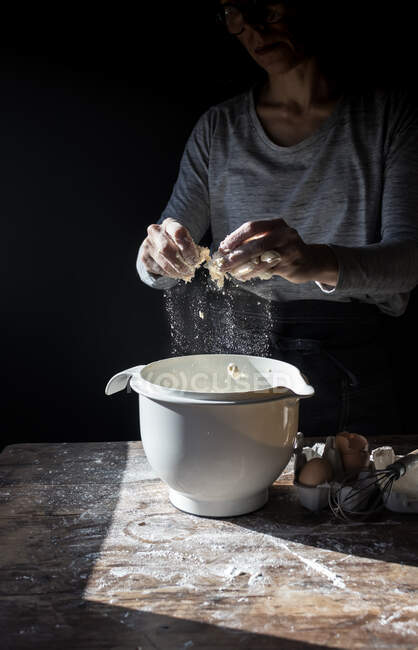 Crop human breaking egg in bowl on wooden table with flour on black background — Stock Photo