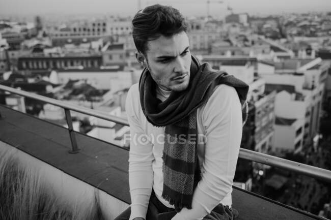 Black and white young stylish guy in casual wear with scarf sitting on roof and looking away on cityscape background in Madrid, Spain — Stock Photo