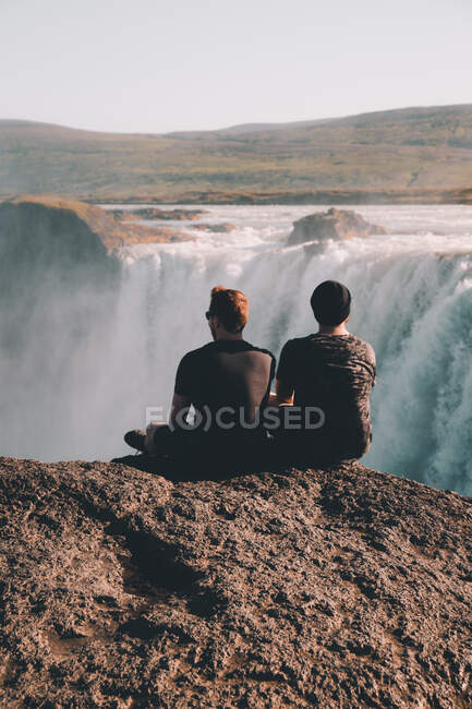 Back view of two men sitting on cliff and admiring amazing waterfall in Arctic countryside - foto de stock