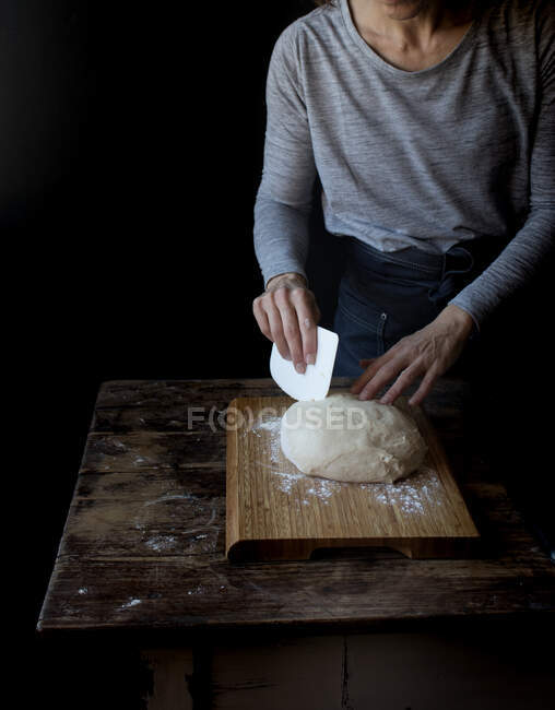 Обрізати людину, що тримає тісто з борошном на подрібненій дошці біля дерев'яного столу на чорному тлі — стокове фото