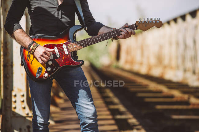 Crop Adult guy with electric guitar standing on weathered bridge on sunny day in countryside — Stock Photo