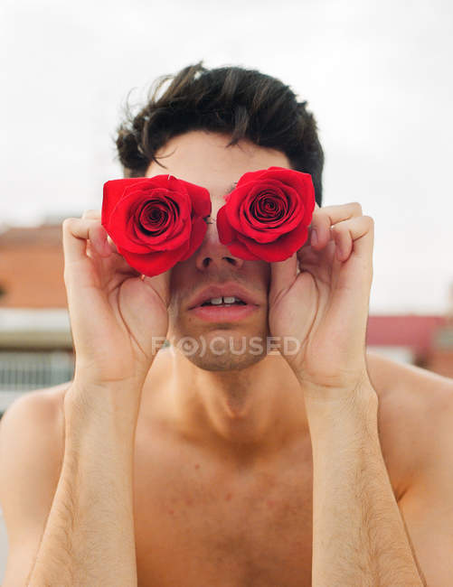 Brunette jeune homme torse nu montrant des roses fraîches vineuses couvrant les yeux sur fond flou — Photo de stock
