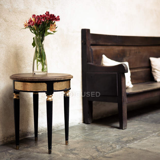 Vase with amazing flowers standing on vintage table near wooden bench inside old building — Stock Photo