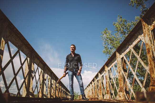 Hombre adulto con la guitarra eléctrica de pie en el puente resistido y mirando hacia otro lado en el día soleado en el campo - foto de stock