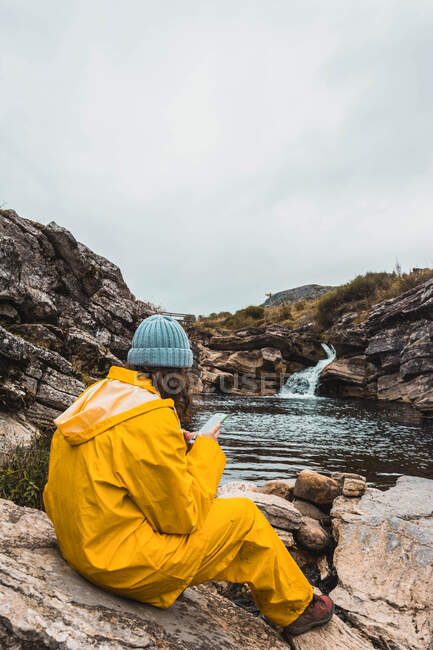 Visão traseira de senhora de chapéu e capa de chuva amarela sentada em pedras fenda de rio entre colinas de rocha usando telefone celular em Isoba, Castela e Leão, Espanha — Fotografia de Stock