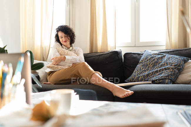 Жінка з подушкою на дивані біля столу з щітками та ноутбуком. — стокове фото