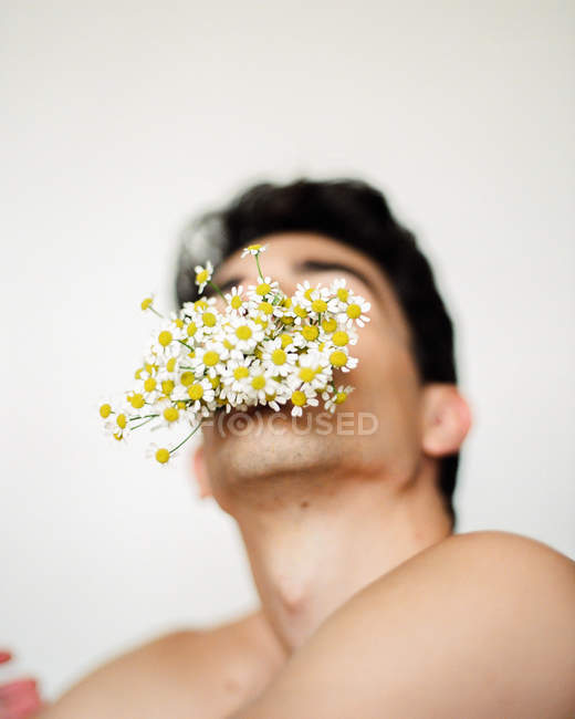 Chico joven sin camisa con flores blancas frescas en la boca mirando a la cámara en un fondo borroso - foto de stock