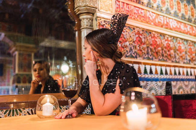 Attrayant jeune femme en robe assis à la table près du verre de boisson et des bougies allumées dans la chambre de luxe du café décoré par la mosaïque — Photo de stock