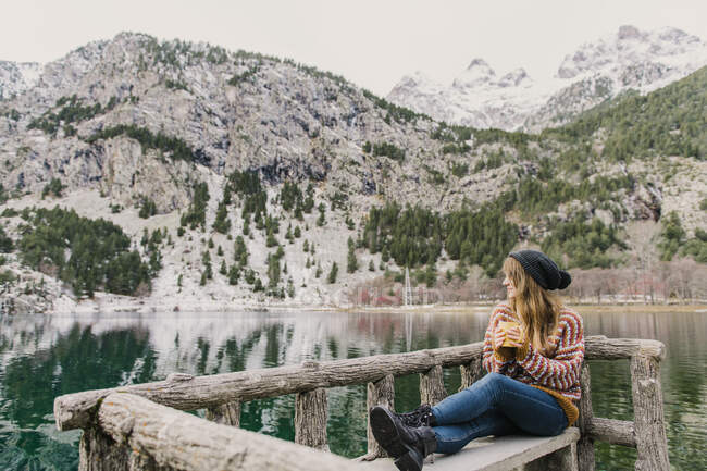 Joven dama sentada en el banco y mirando hacia otro lado cerca de la increíble vista de la superficie del agua entre altas montañas con árboles en la nieve en los Pirineos - foto de stock